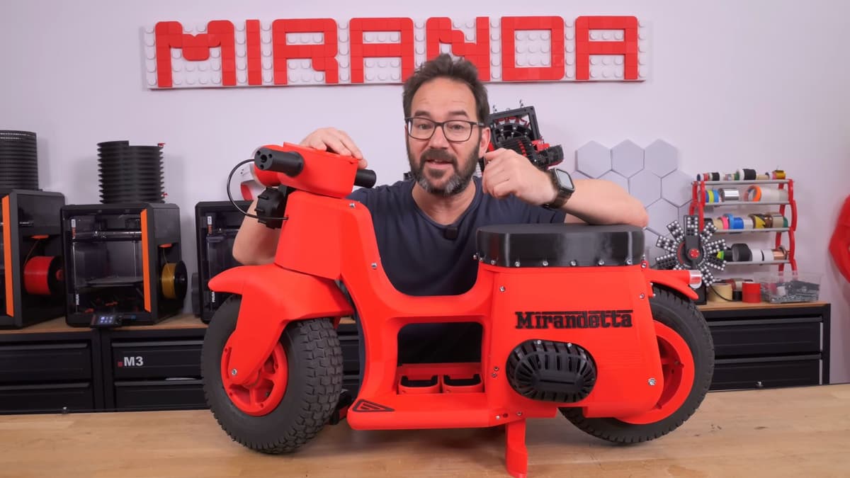 Man smiling next to red Mirandetta motorbike with 3D printers behind